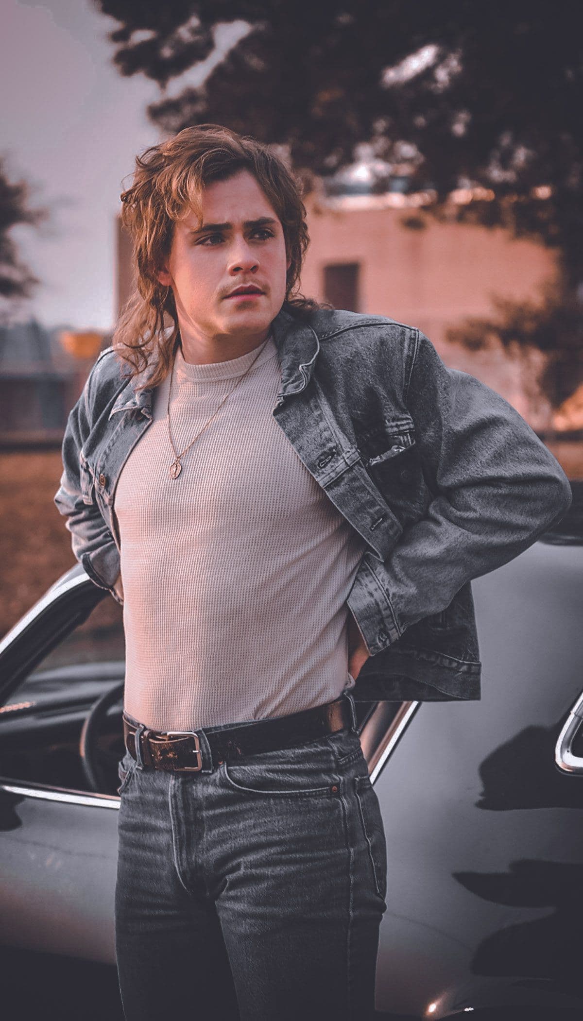 Billy Hargrove aus Stranger Things in schwarzer Jeansjacke und hellem Shirt neben einem Auto.